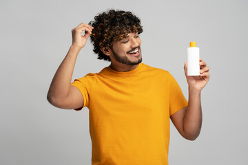 Smiling curly haired Indian man holding bottle with hair shampoo, looking at cosmetic product isolated on gray background, studio shot. Hair care concept  