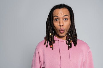shocked multiracial man in pink hoodie pouting lips and looking at camera isolated on gray
