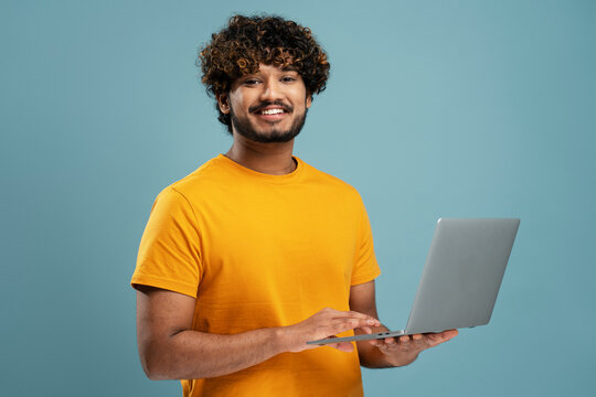Smiling Indian man holding laptop computer working online looking at camera. Happy confident university student studying isolated on yellow background, online education. Portrait of happy programmer  