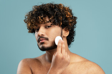 Closeup portrait of attractive metrosexual Indian man removing makeup using cotton disk, cleaning face, looking at mirror isolated on blue background. Beauty, skin care, morning routine concept  