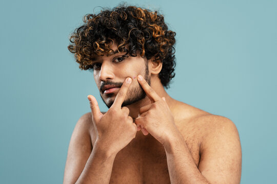Closeup portrait of young serious Indian man removing pimple or acne, cleaning face, looking at mirror isolated on blue background. Beauty, skin care, morning routine concept