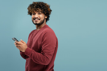 Young  smiling Indian man holding smartphone using mobile app shopping online isolated on blue background, copy space. Portrait of happy asian hipster with curly hair ordering food on website