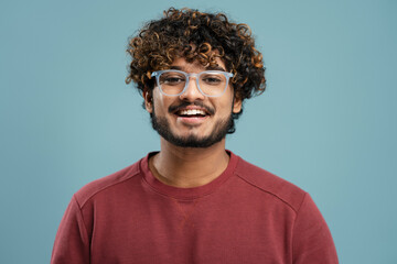 Portrait of attractive smiling curly haired Indian man wearing stylish eyeglasses looking at camera isolated on blue background. Vision concept