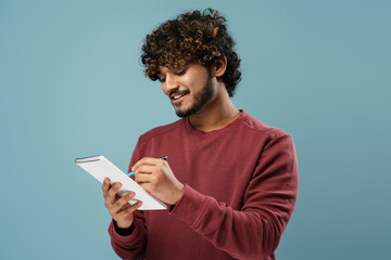 Portrait of smiling Indian man taking notes in notepad isolated on blue background. Smart asian university student studying, learning language, exam preparation. Education concept 