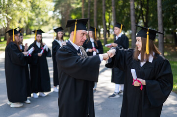 A group of graduates in robes outdoors. An elderly man and a young woman congratulate each other on their graduation.