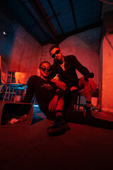 a couple of gay teenagers are sitting together wearing sunglasses and posing naughty in a warehouse with a red light