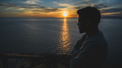 Fototapeta premium Hombre mirando el mar y el océano con el sol en el horizonte al atardecer, colores y nubes en el cielo