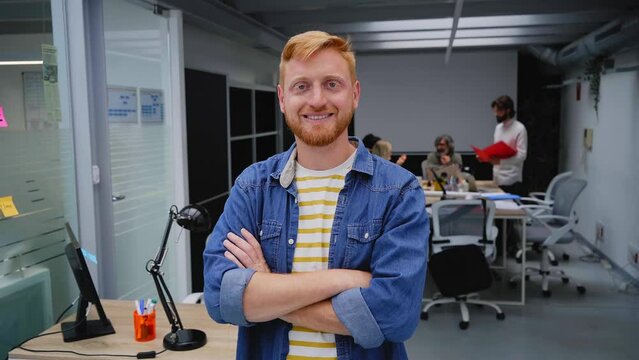 Young red-haired programmer or businessman in a modern office looking camera and smiles. Self-employed guy with his arms crossed poses for a video in the meeting room. Colleagues work in background.