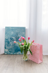 A bouquet of Pink tulips in a glass vase, pink gift bag, turquoise modern painting stand in a room. Background for Valentine's Day. Gift for Mother's Day, Women's Day, March 8th. Spring background