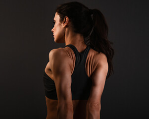 Serious female sporty muscular with ponytail doing stretching workout the shoulders, blades and arms in sport bra, standing on dark grey background with empty copy space. Back view. Lifestyle