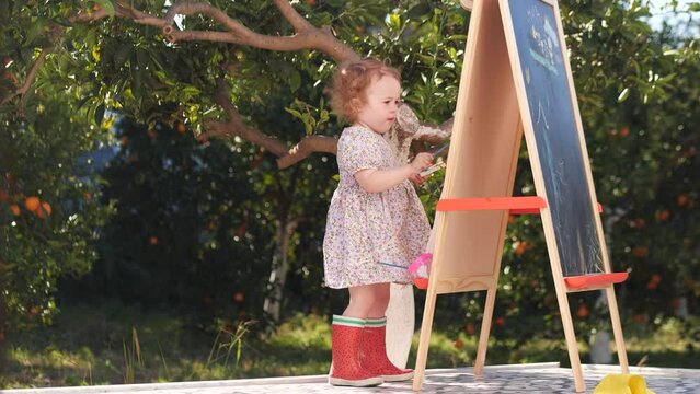 Little baby girl wearing red rain boots paints on easel in orange garden