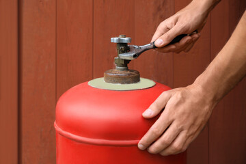 Man with adjustable wrench opening red gas cylinder near brown wooden wall, closeup