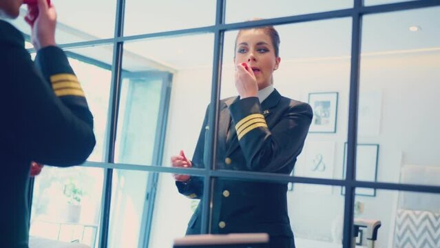 Slow motion of serious female pilot applying lipstick and looking in mirror, confident young adult pilot in uniform and looking in mirror standing near suitcase in hotel room before flight