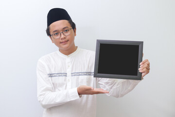 Portrait of young Asian muslim man holding a blackboard with hand pointed. Blank space for typography or text to fill. Isolated image on white background