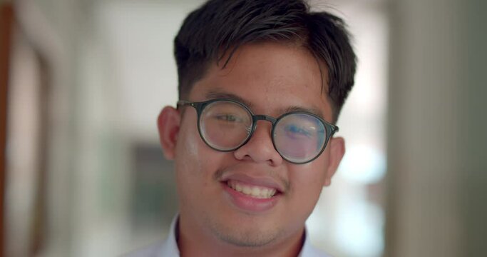 A little smile of an Asian male teenage high school student wearing thick glasses standing on the corridor in the building.