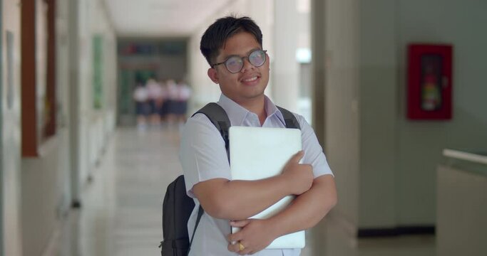  A happy smile of an Asian male teenage high school student wearing thick glasses. wearing a white sweater, carrying a laptop with a backpack standing on the corridor in the building.