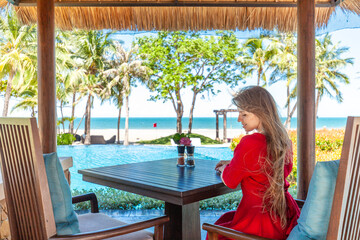 Woman on summer vacation. Travel girl in red dress and hat sitting on breakfast at the table in hotel lounge near pool. Tropical beach restaurant with sea view.