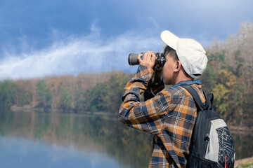 Asian boy in plaid shirt wears cap and has a backpack, holding a binoculars, standing on ridge reservoir in local national park to observe fish and birds on tree branches and on sky, soft focus.