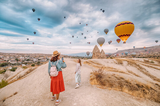 Two girls friends capture the beauty of Cappadocia, Turkey, with their smartphones amid stunning rock formations and hot air balloons.