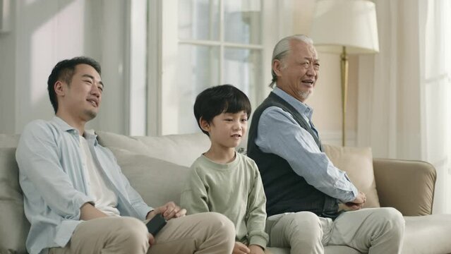 three generation asian sports fans son father grandfather sitting on couch watching soccer match on tv