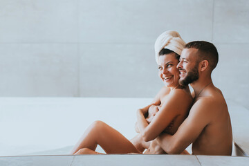 Young happy couple sits in bath naked laughing enjoying honeymoon at hotel. Beard caucasian guy hugs beloved wife at morning, bathroom. Pretty hispanic girl with towel on head cuddling with boyfriend.