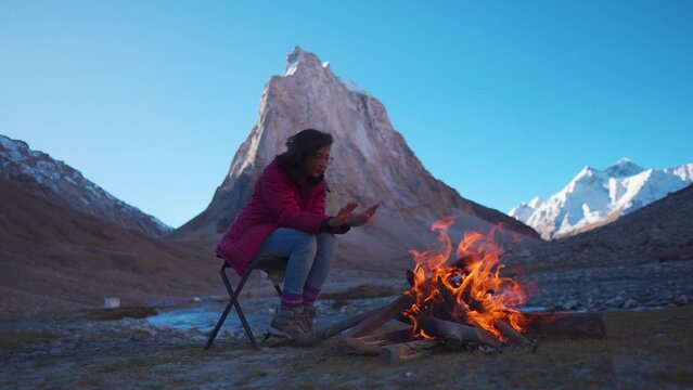 4K shot of young Indian woman sitting on chair with fire at mountain during cold winter morning at Zanskar, Ladakh, India. Girl sitting near a fireplace on background of Holy Gonbo Rangjon mountain. 