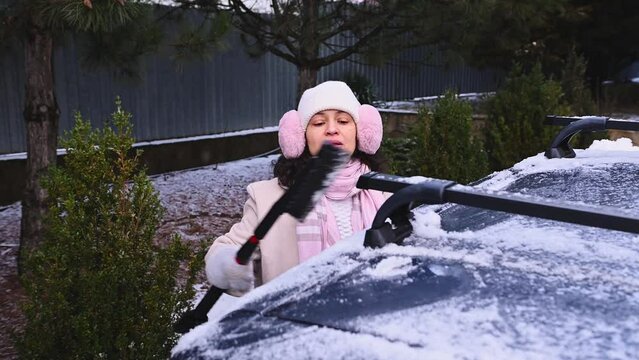 Woman using automobile brush sweeping off snow from her car during winter snowfall. Winter scene. Driver scraping snow from vehicle. Daytime. Transportation, winter, weather, people, vehicle concept