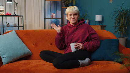 Obraz premium Young woman with short blonde hair smiling friendly at camera and waving hands gesturing hello, hi, greeting or goodbye, welcoming with hospitable expression. Girl indoors at home in room on couch