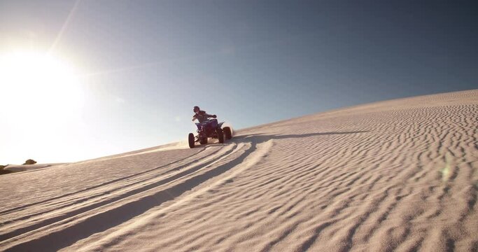 Competitive quad bike racer kicking up sand while driving up a sand dune on a summer evening with sun flare, Slow Motion, Panning