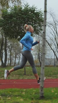Vertical Screen: Slow motion fit woman wearing blue sports jacket running on track in autumn park, fallen leaves lying on ground. Tracking shot of active person training in morning. Concept of fitness