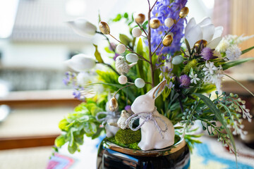 Easter table decoration made ot artificial flowers, green leaves, birds, and small eggs. White glass Easter bunny sitting in flower pot.