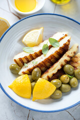 Slices of grilled haloumi cheese with lemon and green olives in a white plate, closeup, vertical shot