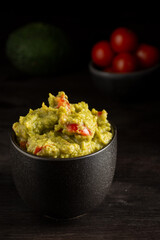 Top view of black bowl with guacamole on dark table with small tomatoes and avocados, selective focus, vertical with copy space