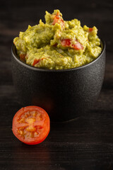 Top view of black bowl with guacamole on dark table with half a small tomato, vertical with copy space