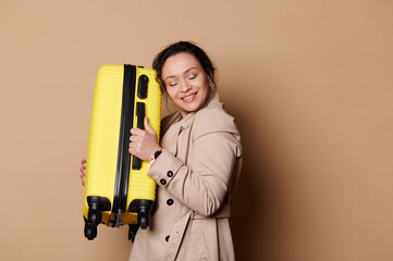Delightful Caucasian woman, wearing a stylish beige coat, hugging her yellow suitcase, smiling a beautiful toothy smile, posing with her eyes closed, over beige background. People and Travel concept