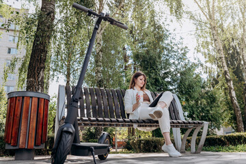 Pretty business woman in a white suit sits in a city park, drinks coffee and works on a digital tablet with an electric scooter in the foreground