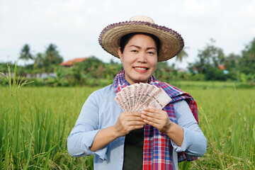 Fototapeta premium Asian farmer woman is at paddy field, wears hat and red plaid shirt, hold Thai banknote money. Concept : Farmer happy to get profit, income, agriculture supporting money. 