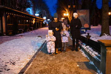 Obraz premium Family walk through the night city in the winter. A woman and three children in evening drink hot drink in cups.