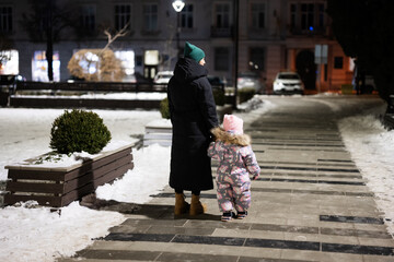 Back of mother walk through the night city in the winter. Mom and daughter in evening.