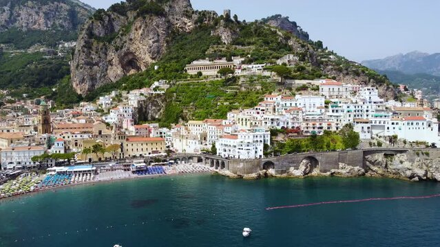 Aerial drone footage of the Marina Grande beach, town hall, Sant'Andrea duomo bell tower in Amalfi coast, Campania, Italy. Parasol, sun loungers, people swimming and colourful cliff villas from above