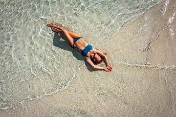 A young woman in a blue bikini lies on her back on the sand near the waves of the blue sea. View...