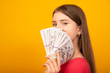 Young woman holds a stack of dollar bills fanned near her face against a bright yellow orange background. Copy space