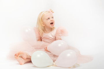 Laughing blonde girl in pink lush dress with balloons sits on floor. Happy child on her birthday. White background