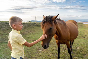 Obraz premium Teenage boy strokes horse in the field outdoor against background of hills and mountains at sunset. Boy taking care of broen horse on meadow in wild nature