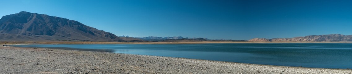 Panoramic view of Crowley Lake and the surrounding mountains, during a sunny day. A person walks near the edge of the lake.