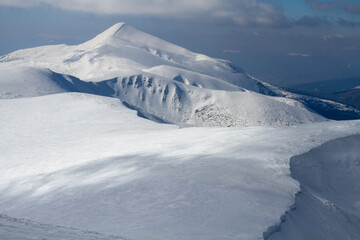 A large white rocky mountain with a dark couloir is covered with snow among the foggy clouds before winter blizzard
