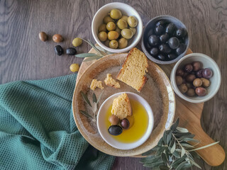 Table view consisting of several bowls with different types of olives, plate with cornbread and extra virgin olive oil, dark wood table background and olive branches