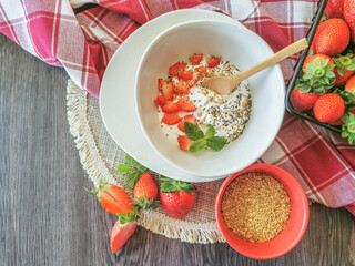 Breakfast view, with a bowl of natural yogurt with pieces of strawberry, mint, linseed and chia seeds; background with whole strawberries, bowl with linseed and kitchen towels on dark wooden table