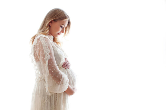 Beautiful pregnant woman with long blonde hair in white dress stands sideways and touches her belly isolated on white background, backlit
