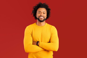 Handsome smiling African American curly young man in casual wear posing on red background, charismatic guy in yellow pullover with toothy smile stand with arms crossed and look at the camera
